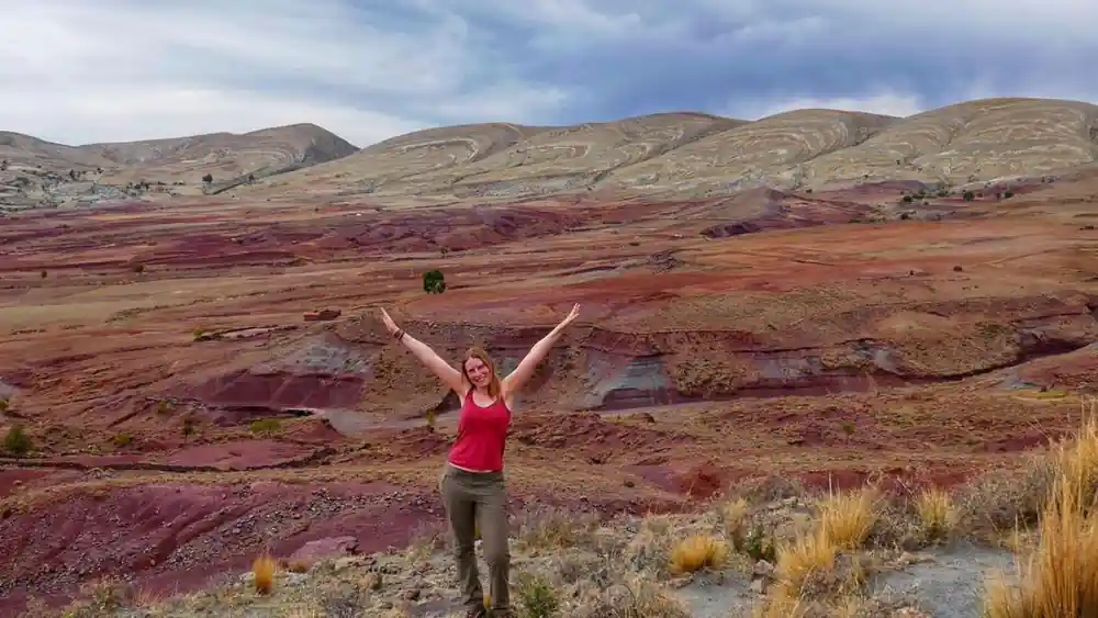 A visitor with arms raised in front of the red and ochre bands of the crater